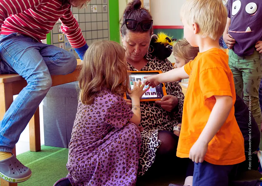 Erzieherin, die ein Tablet in der Hand hält und alle Kinder um sie herumstehen und auf das Tablet schauen
