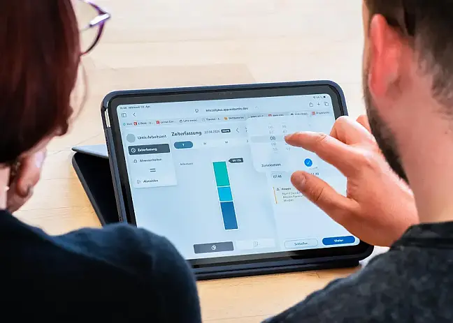 Ein Tablet, an dem zwei Lehrkräfte sitzen und das Arbeitszeitprogramm zu sehen ist