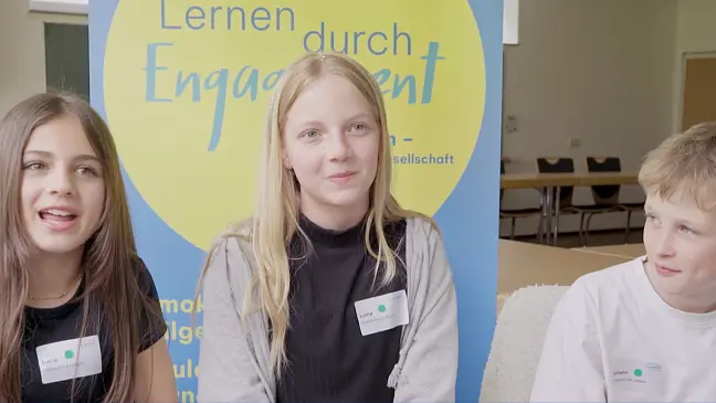 Zwei Schüler:innen und ein Schüler, die in einer Reihe auf einem Stuhl sitzen. 