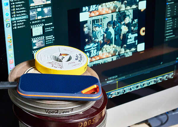 Filmrollen stapeln sich vor einem Monitor, der in einem Videoschnittprogramm ein Filmprojekt zeigt.