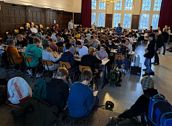 Schulschach. Viele Schüler:innen sitzen an Tischen und spielen Schach. 